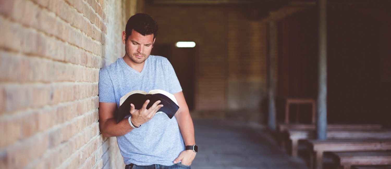 Man standing against brick wall reading Bible